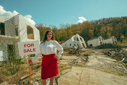 Smiling realtor holding for sale sign by destroyed house outdoors