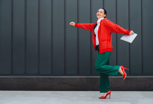 Happy freelancer jumping with laptop outdoors in red cardigan