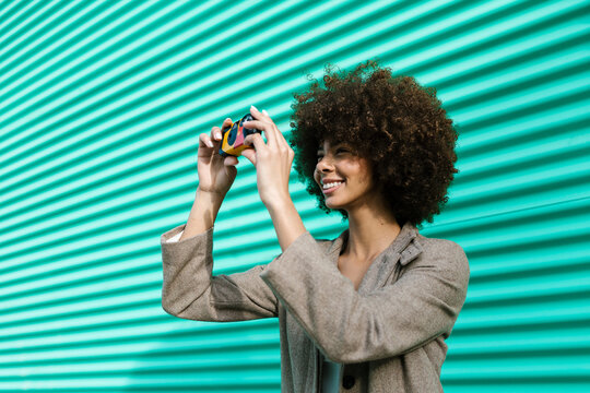 Cheerful woman with afro taking photo against turquoise wall