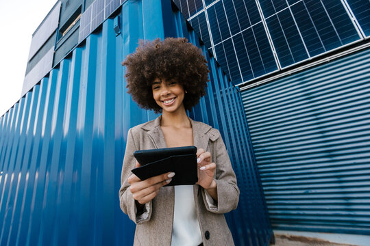 Cheerful female entrepreneur with tablet near container and solar panel