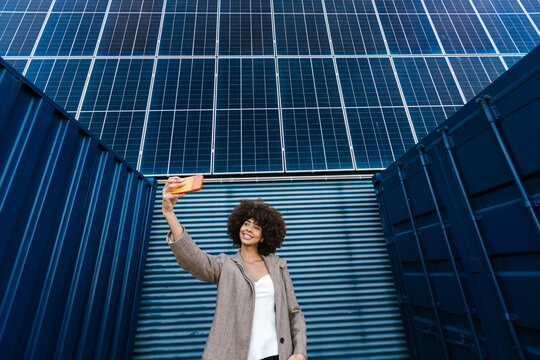 Woman taking a cheerful selfie with a smartphone near a solar panel