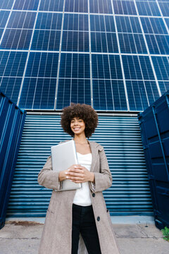 Smiling businesswoman holding laptop by solar panel outdoors