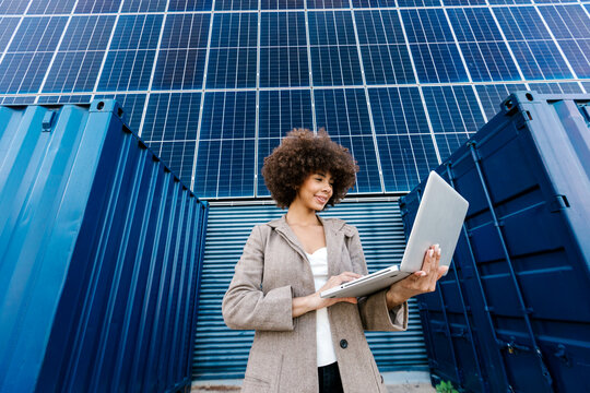 Female engineer using laptop near solar panels and shipping containers