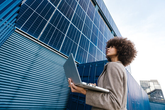 Female engineer holding laptop looking up near solar panel outdoors
