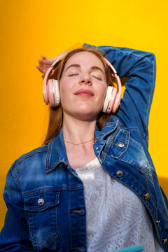 Redhead listening to music with headphones against yellow wall