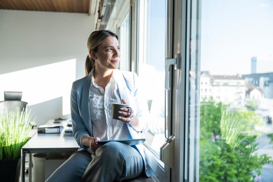 Smiling entrepreneur sitting by office window holding tablet and mug