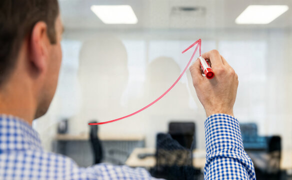 Businessman drawing upward trending arrow on glass wall in office with chairs and tables in background.