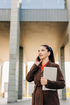Smiling businesswoman holding laptop and talking on smartphone