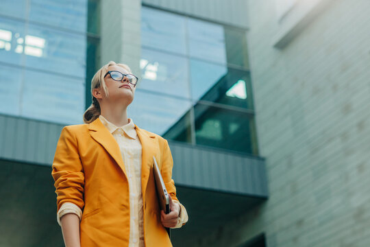 Businesswoman in yellow blazer walking outdoors with laptop