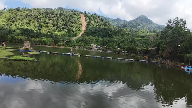 Aerial view of Rawa Gede Jonggol Lake featuring a floating bridge walkway surrounded by dense tropical forest and misty mountains in Kecamatan Sukamakmur, Jawa Barat, Indonesia.