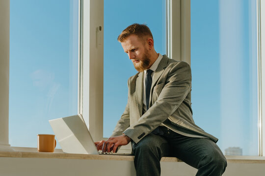 Businessman in suit sits on office windowsill working on laptop