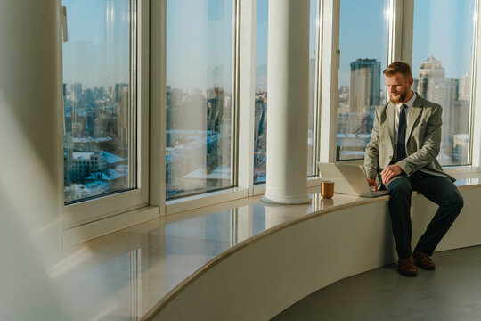 Businessman working on laptop sitting on office windowsill