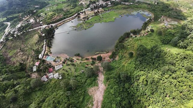 Aerial view of Rawa Gede Jonggol Lake surrounded by lush green hills and rural settlements in Kecamatan Sukamakmur, Jawa Barat, Indonesia.
