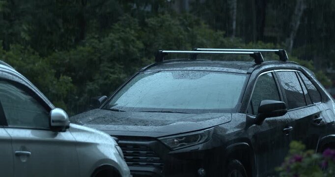 Close-up of a wet crossover vehicle with water droplets on its windshield and hood, parked in a dim, moody evening setting near trees.