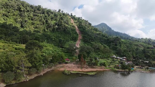Aerial view of Rawa Gede Jonggol Lake surrounded by lush green hills and a visible landslide on the mountain slope in Kecamatan Sukamakmur, Jawa Barat, Indonesia.