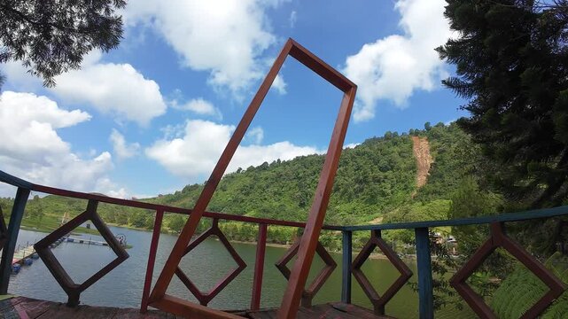 Aerial view of Rawa Gede Jonggol Lake from a wooden deck with geometric railings surrounded by lush green hills and a blue sky in Kecamatan Sukamakmur, Jawa Barat, Indonesia.