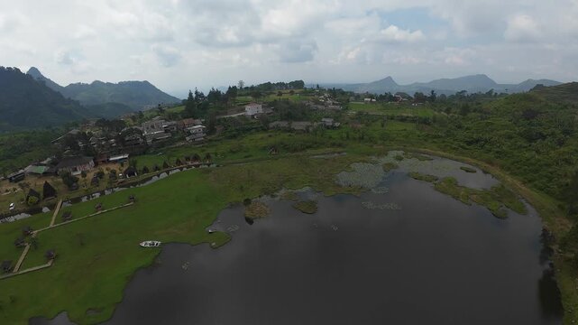 Aerial view of Rawa Gede Jonggol Lake surrounded by lush green hills, rural houses, and terraced fields under a cloudy sky in Kecamatan Sukamakmur, Jawa Barat, Indonesia.