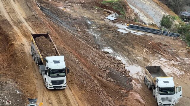 Heavy dump trucks transporting soil on a steep, muddy construction site [cite: MMM.jpg]. High-angle operation shot of infrastructure, excavation, and earthworks