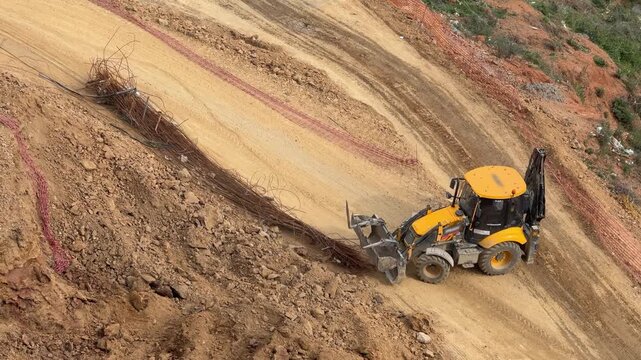 Backhoe loader handling steel rebars and working on a steep slope at a construction site [cite: MM.jpg]. Infrastructure development and slope stabilization field operation