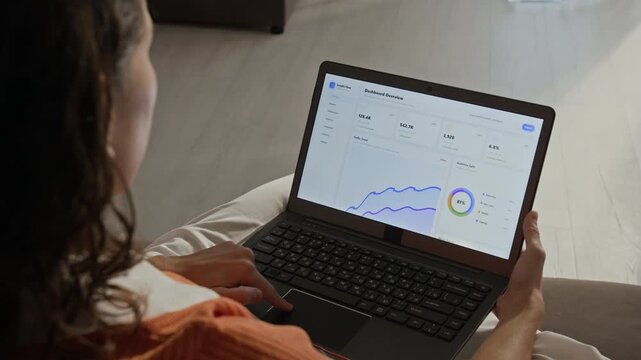 Over shoulder shot of Caucasian young adult woman analyzing business metrics and scrolling through dashboard analytics on laptop. Clip perfect for marketing campaigns about remote work and e-commerce