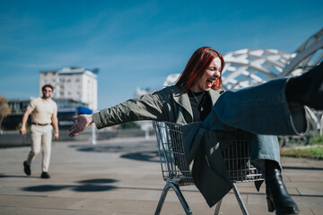 Plakat A young red-haired woman laughs while sitting in a shopping cart outdoors. Her friend walks toward her as they enjoy a playful moment in the city