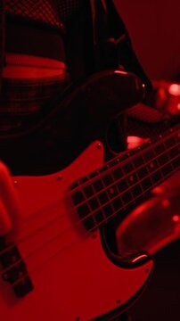 Vertical video of young Caucasian female musician playing electric bass guitar during band rehearsal under red light. Footage is suitable for music festival promos or concert advertisements