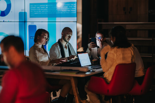 Business teammates work together around a table with laptops and charts on a large screen. A small team collaborates on data analysis during a focused late-night office session