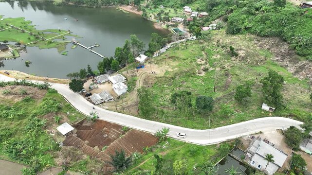 Aerial view of Rawa Gede Jonggol Lake with a winding road, lush green vegetation, and small buildings on the hillside Kecamatan Sukamakmur, Jawa Barat, Indonesia.