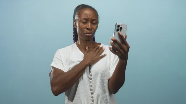 African american woman holds smartphone and clutches chest in studio, looking concerned; vulnerability concern.