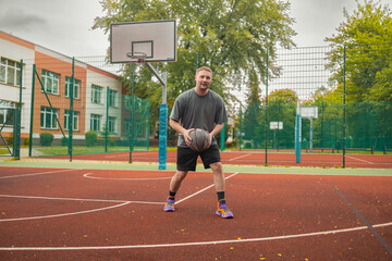 Fototapeta premium Full length portrait of a young smiling man holding a basketball ball during training on an outdoor playground