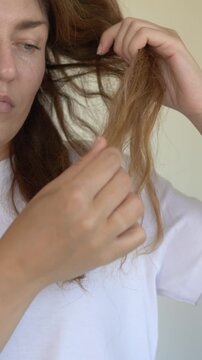 Woman pulls and examines damaged hair strand checking breakage loss and split ends in haircare concern closeup. Footage suits anti-breakage shampoo brand, trichology clinic content, hair loss beauty