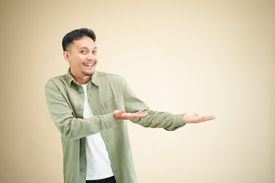Happy young Asian man presenting with one open palm extended sideways toward copy space and smiling, casual olive green shirt in beige studio portrait
