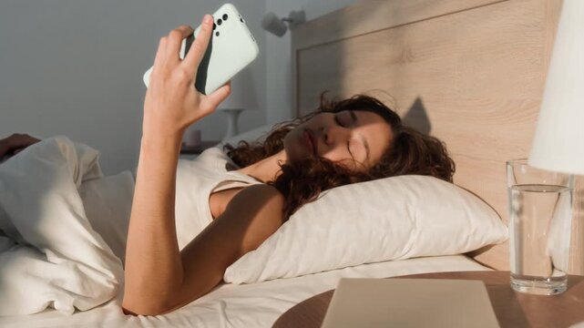 Asian young adult woman waking up in bed, reaching for smartphone on nightstand and scrolling. Video is ideal for mobile application ads, morning routine promos or social media campaigns