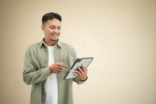 Young Asian man tapping on a digital tablet screen while smiling, using the device in casual olive green shirt against beige studio background