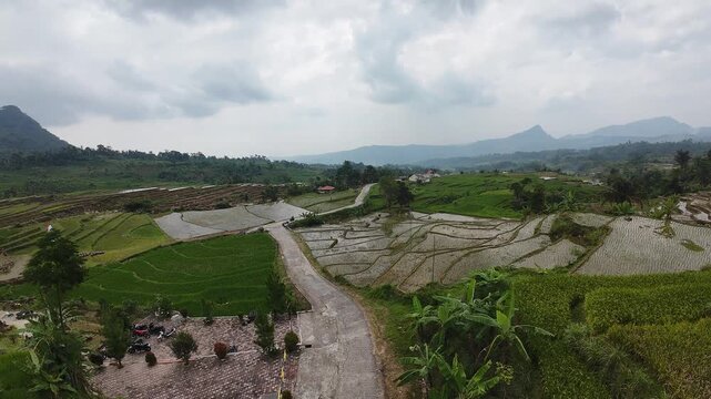 Aerial view of lush green rice terraces and a winding road near Rawa Gede Jonggol Lake under a cloudy sky in Kecamatan Sukamakmur, Jawa Barat, Indonesia.