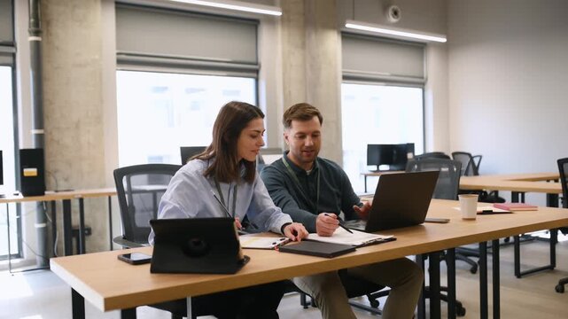 Two office workers, a woman and a man, collaborate at a desk with a laptop and documents in a contemporary workspace setting