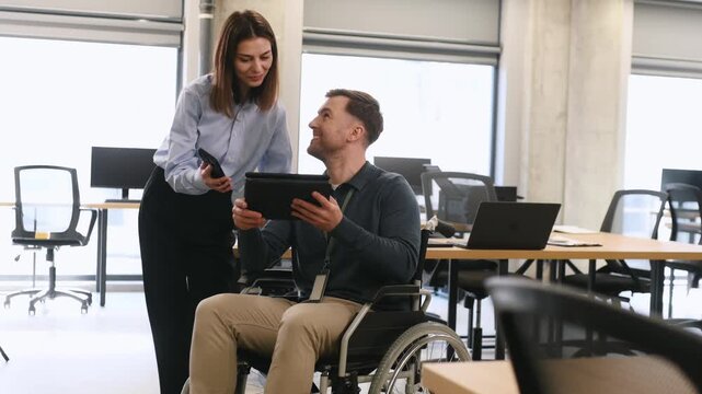Businesswoman interacts with a man in a wheelchair using a laptop in a contemporary office environment featuring large windows and wooden furniture