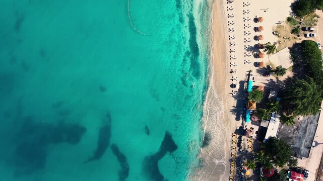 Aerial view of Saint Martin island in the Caribbean Sea. Clear turquoise water of the sea next to a sandy beach. Rows of umbrellas and sunbeds with people on the coast. 