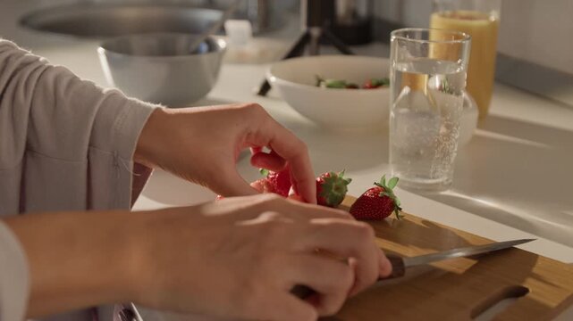 Young adult Caucasian woman slicing fresh strawberries on wooden cutting board in kitchen, preparing healthy breakfast. Footage is perfect for wellness campaigns or healthy eating promotions