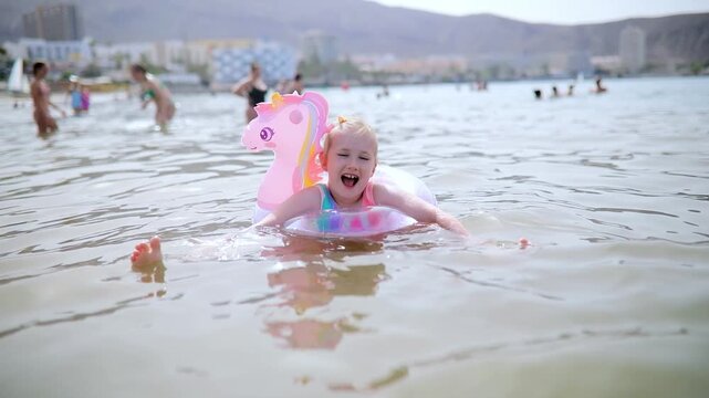Little girl playing in shallow sea water with an inflatable unicorn swim ring in Los Cristianos, Tenerife, Canary Islands, Spain