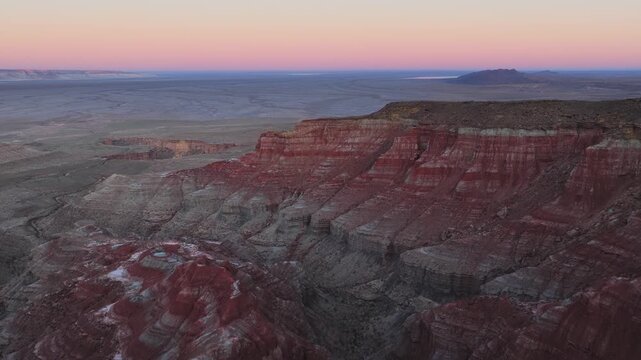 Aerial view of the red layered cliffs of Karynzharyk hollow under a soft pink sunset sky in Karakiya District, Mangystau Region, Kazakhstan.