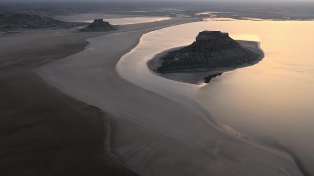 Aerial view of the Karynzharyk hollow featuring rocky buttes rising from the vast salt flat under a golden sunset sky in Karakiya District, Mangystau Region, Kazakhstan.