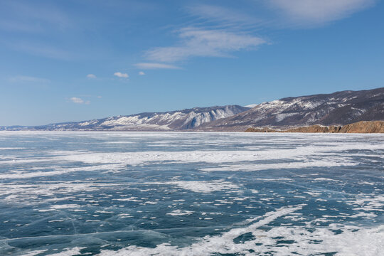 Wide winter view of frozen Lake Baikal with deep blue ice, cracks and snow patterns, and distant mountains under clear sky in Irkutsk Oblast, Russia