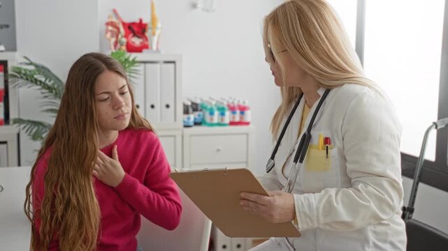 Two women doctor and patient, patient hand on chest while doctor with stethoscope reads clipboard in building; concern medical consultation.