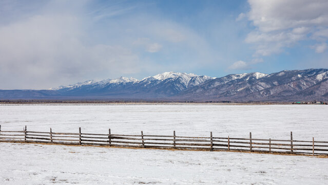 Wide winter landscape of snowy steppe with a rustic wooden fence and distant snowcapped mountain range under cloudy sky in Buryatia, Siberia, Russia