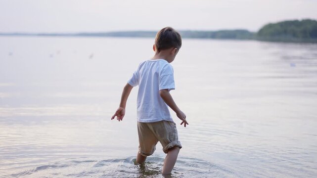 Rear view of a toddler wading into a calm lake. Back of child walking through shallow water with outstretched arms and distant green shore.