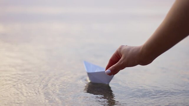 Hand placing a paper boat on water at golden hour. Close up of fingers setting origami vessel on rippled surface with warm backlight.