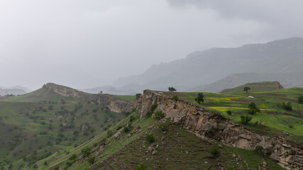 Fototapeta premium Panoramic view of Chokh Terraces with green hills, rocky escarpment and wildflower fields under cloudy sky, mountain scenery in Dagestan, Russia