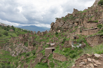 Fototapeta premium Wide view of abandoned stone houses and fortress ruins on a steep green mountainside in Kahib, Dagestan Republic, Russia, under dramatic cloudy sky