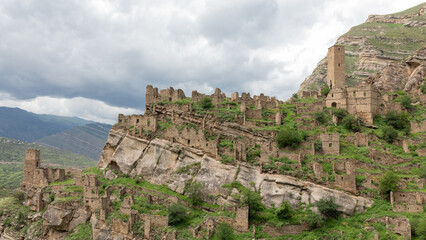 Fototapeta premium Wide landscape view of the abandoned stone village ruins of Kahib on a steep rocky hillside in Dagestan, Russia, with medieval tower houses and cloudy sky